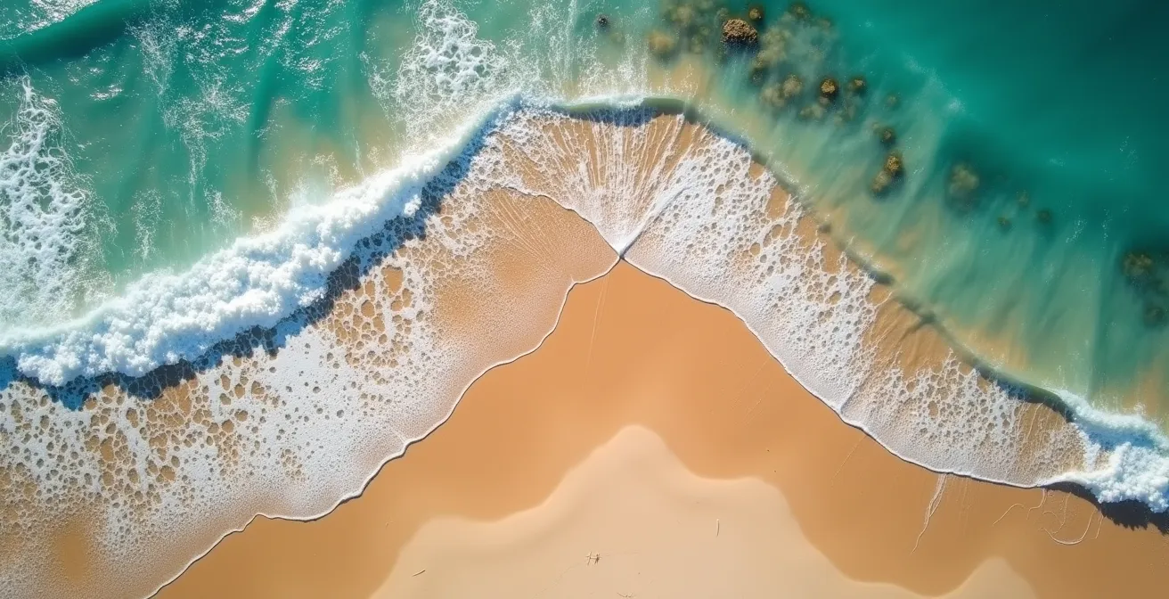 Luftaufnahme eines Sandstrandes, die deutlich einen dunkleren Kanal einer Rippströmung zwischen den brechenden Wellen zeigt